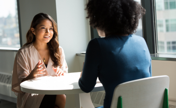 Two women have a conversation at work over a table by the window	